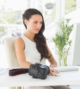 Image of a female photographer wearing a white dress sitting at a computer with a camera.